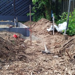 Brukarstwo i Ogrodnictwo Paweł Skarwecki - Przygotowanie terenu pod budowę, widoczne narzędzia, ziemia, drewniane podpory i częściowo wylany fundament z szarych betonowych bloków.