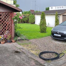 Brukarstwo i Ogrodnictwo Paweł Skarwecki - Zadbany ogród z równo przystrzyżonym trawnikiem, tujami i kwitnącymi kwiatami przy altanie. Na pierwszym planie zwinięty wąż ogrodowy i fragment betonowego podjazdu.