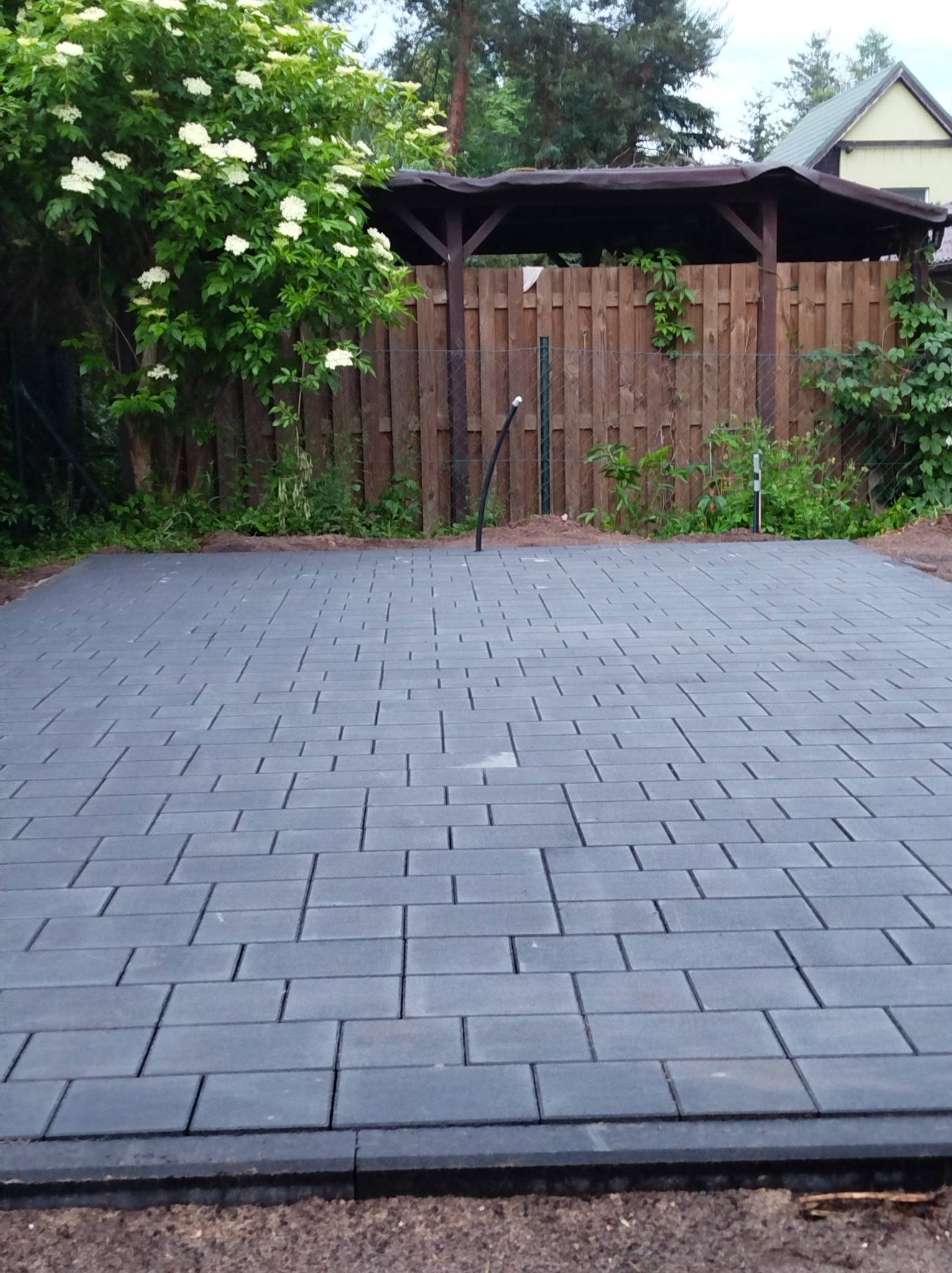 Szara kostka brukowa ułożona na posesji, widoczna zieleń i drewniana altana w tle. Regularny wzór kostki, świeżo zakończone prace.