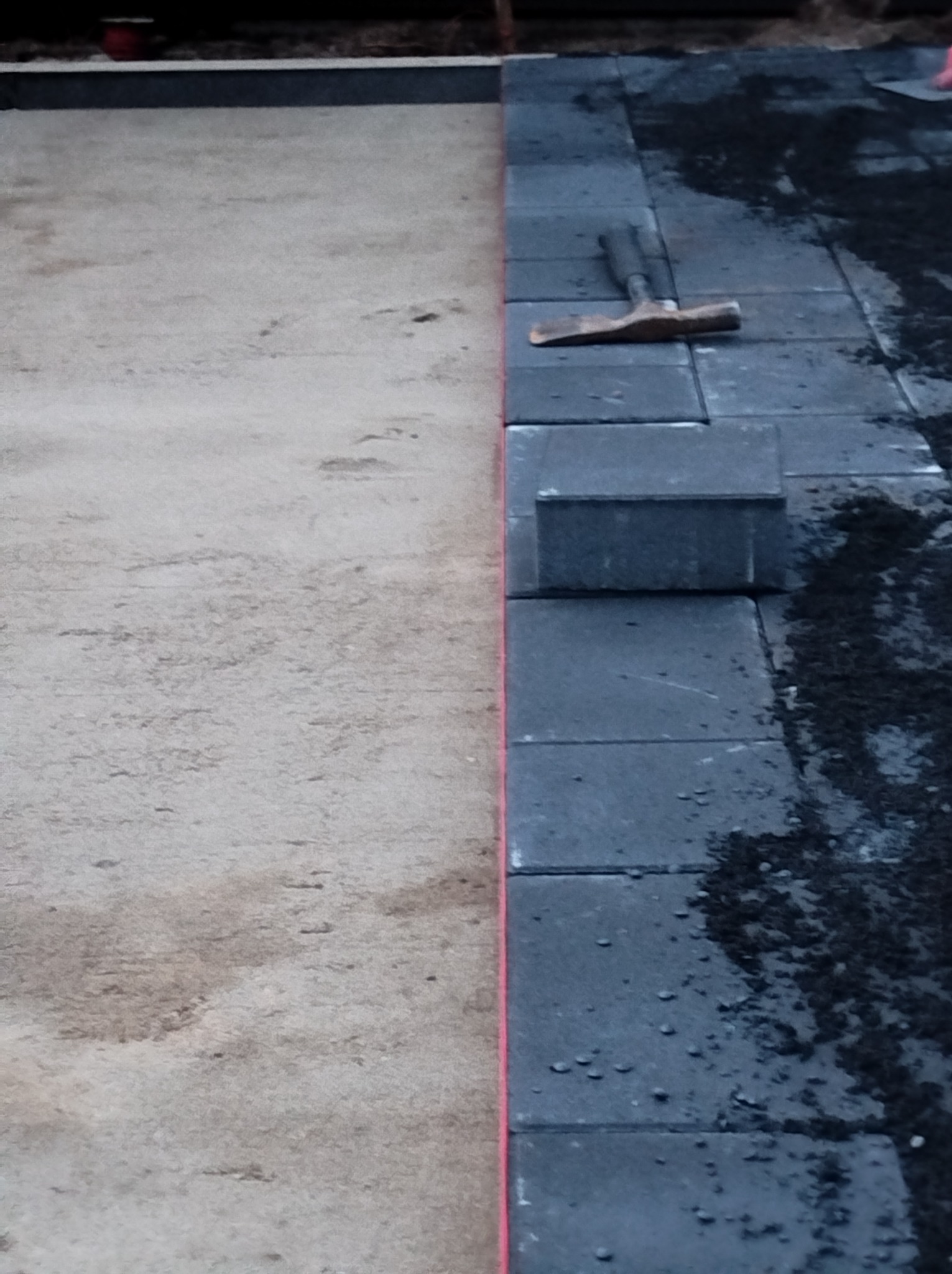 Częściowo ułożona, szara kostka brukowa z gumowym młotkiem, widoczna na tle przygotowanego podłoża z piasku. Układanie w trakcie realizacji.