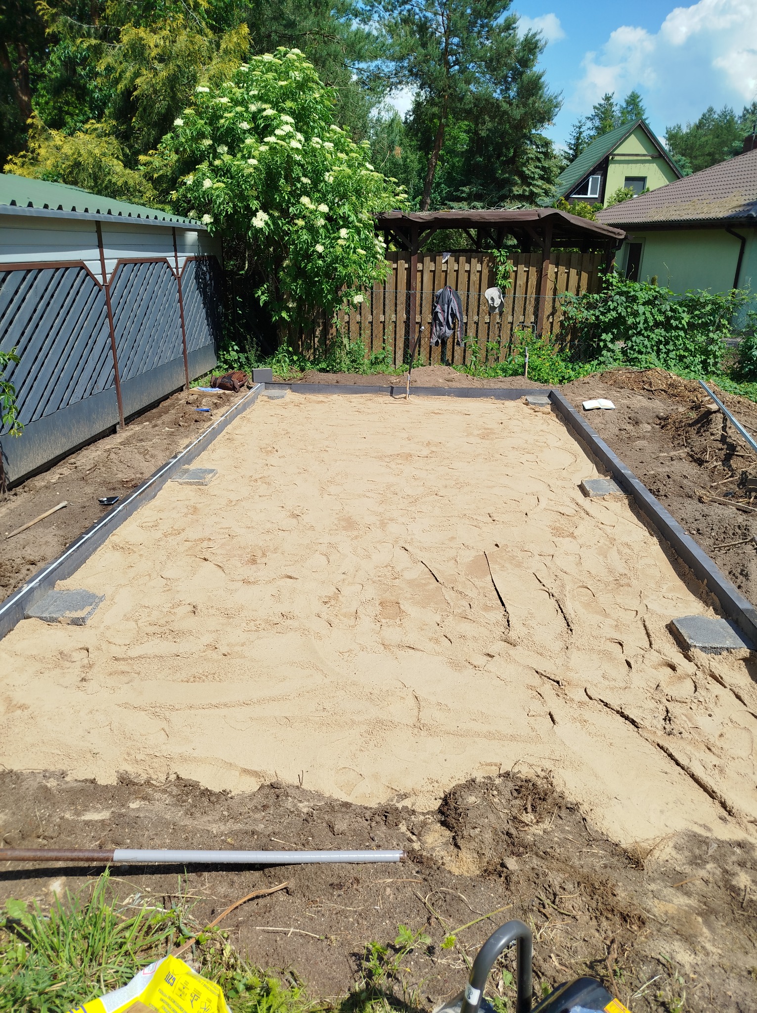 Przygotowany teren pod kostkę brukową z obrzeżami i podsypką piaskową, widok z góry, w tle zieleń ogrodu i zabudowa jednorodzinna.