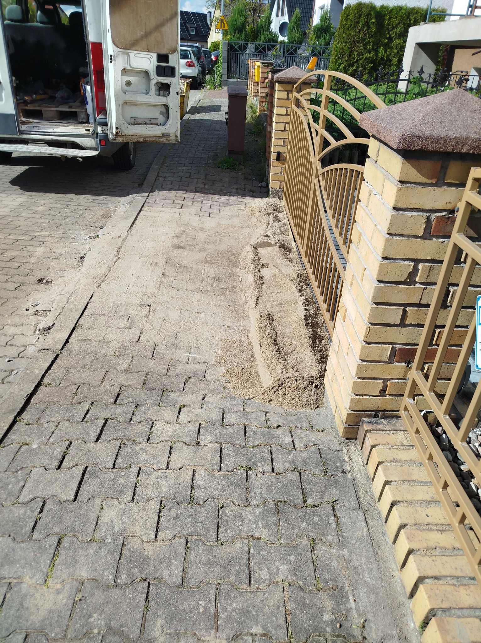Przygotowanie podłoża pod kostkę brukową przy bramie wjazdowej. Widoczny piasek, częściowo ułożona kostka i samochód dostawczy z otwartymi drzwiami.