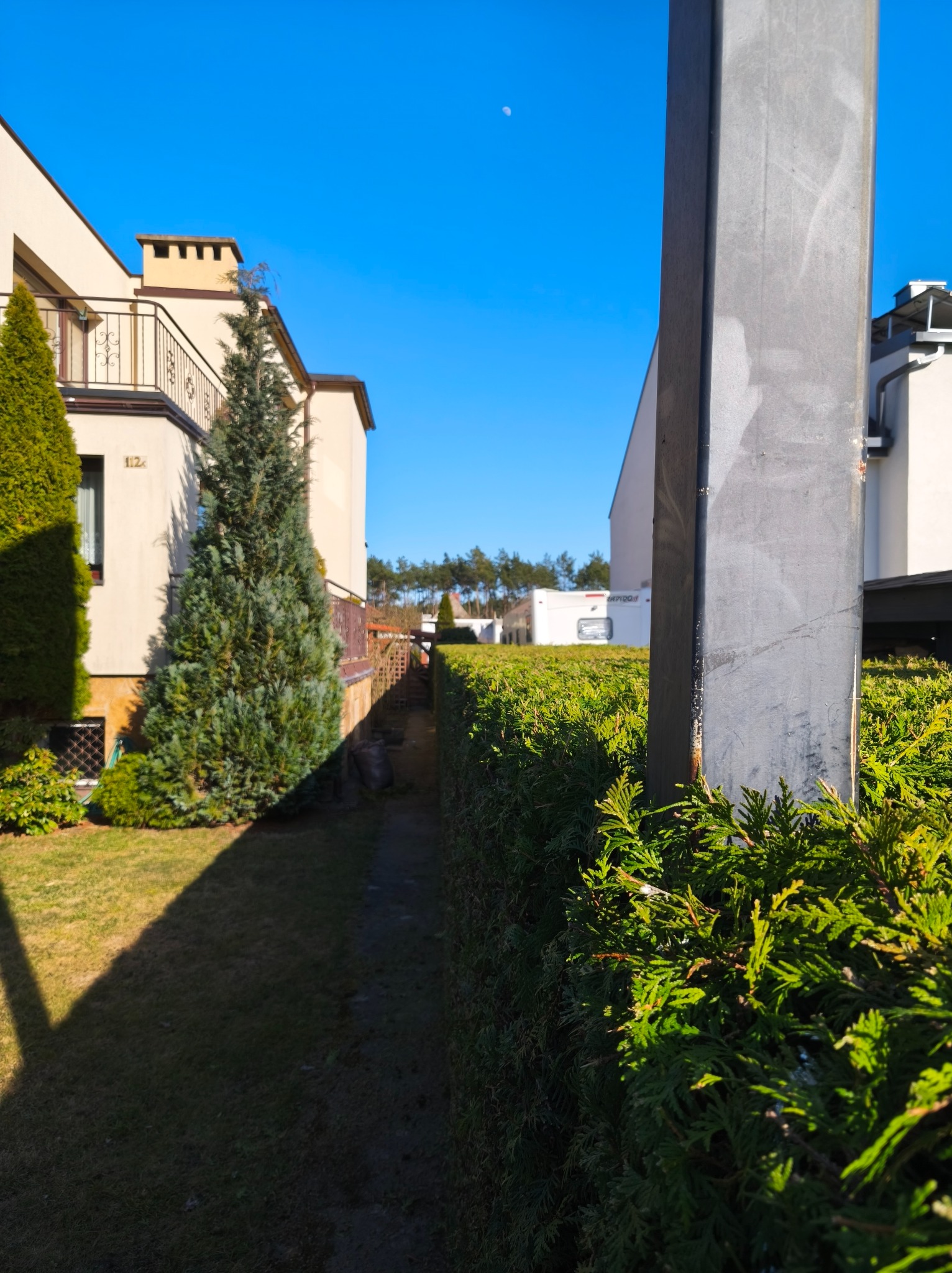 Zadbany ogród z równo przyciętym żywopłotem i strzyżonymi drzewami iglastymi przed domem z balkonem w słoneczny dzień. Widoczny fragment słupa oświetleniowego.