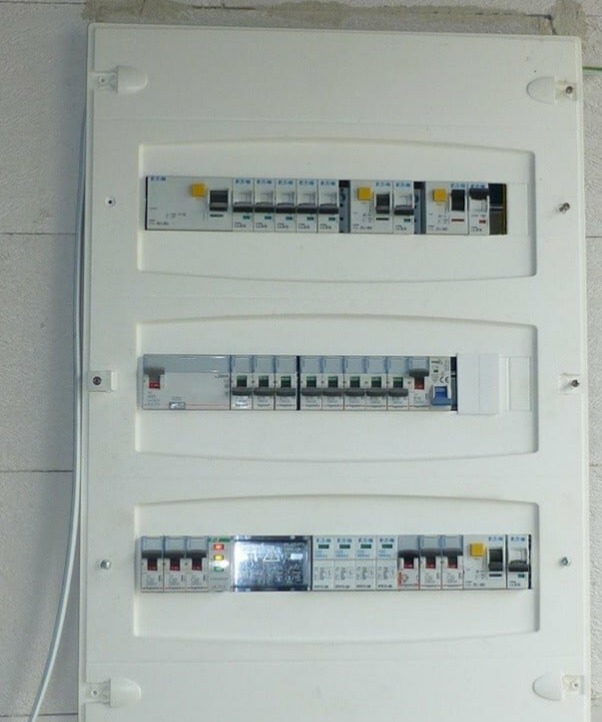 Biała skrzynka rozdzielcza z bezpiecznikami i wyłącznikami, zamontowana na ścianie. Widoczne trzy rzędy aparatury modułowej Eaton.