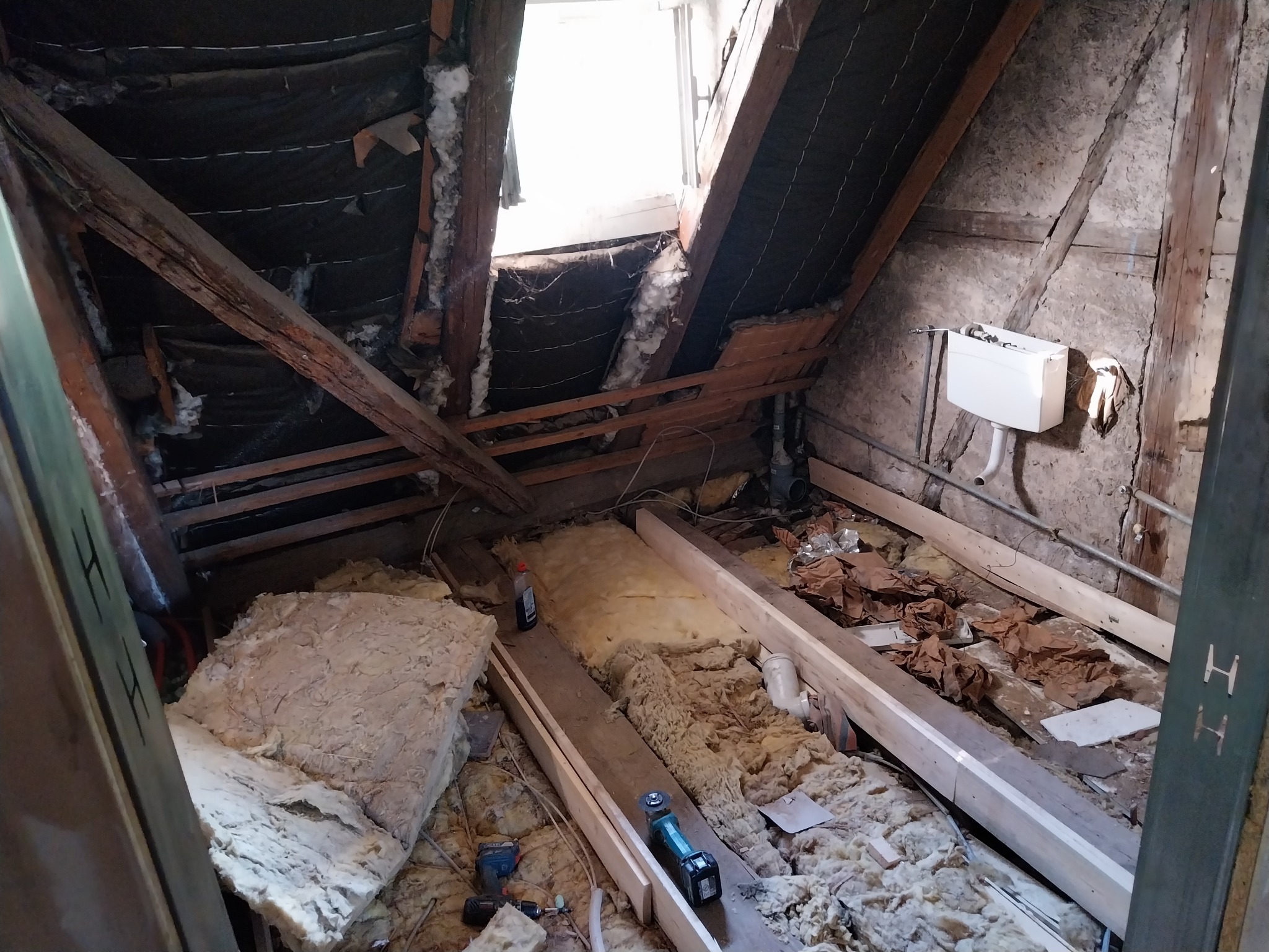 Poddasze w trakcie ocieplania: wełna mineralna, drewniane belki, okno dachowe, instalacja sanitarna i narzędzia robocze w nieuporządkowanym otoczeniu.
