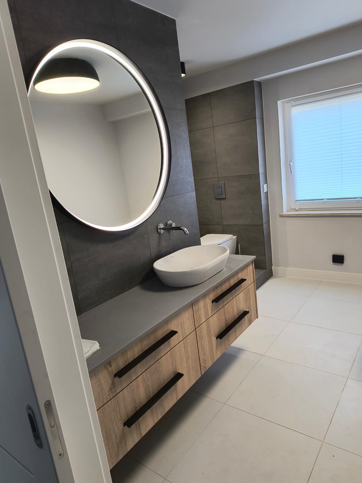 Nowoczesna łazienka z okrągłym lustrem podświetlanym LED, umywalką nablatową na szafce z drewnopodobnymi frontami i szarymi płytkami na ścianach.