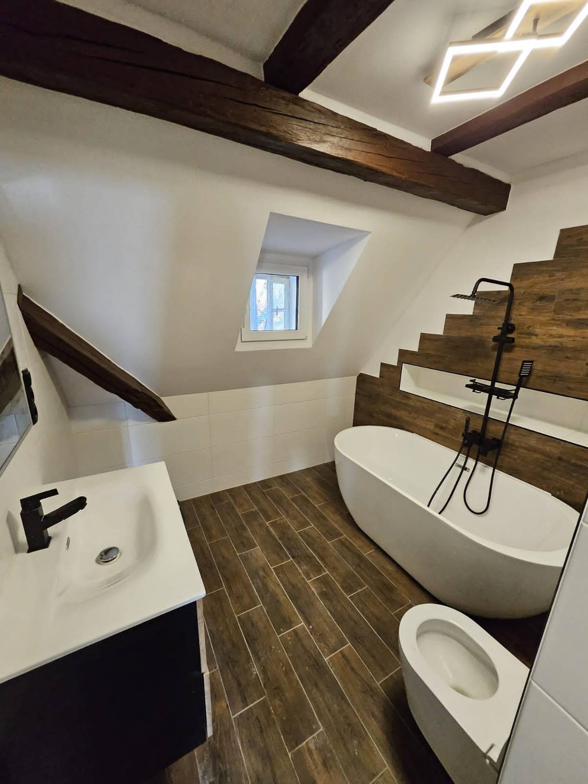 Nowoczesna łazienka na poddaszu z drewnianymi belkami stropowymi, wolnostojącą wanną z czarną armaturą, umywalką z czarną baterią i toaletą.