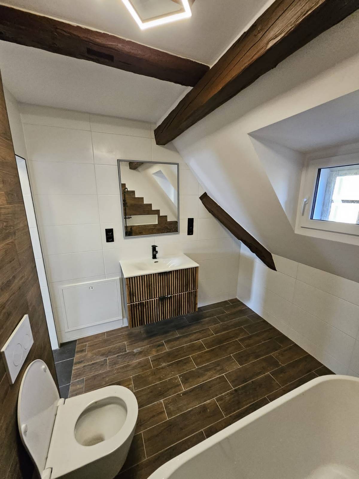 Nowoczesna łazienka na poddaszu z drewnianymi belkami stropowymi, umywalką z szafką z lamelami, lustrem odbijającym schody, białymi płytkami ściennymi oraz toaletą i wanną.