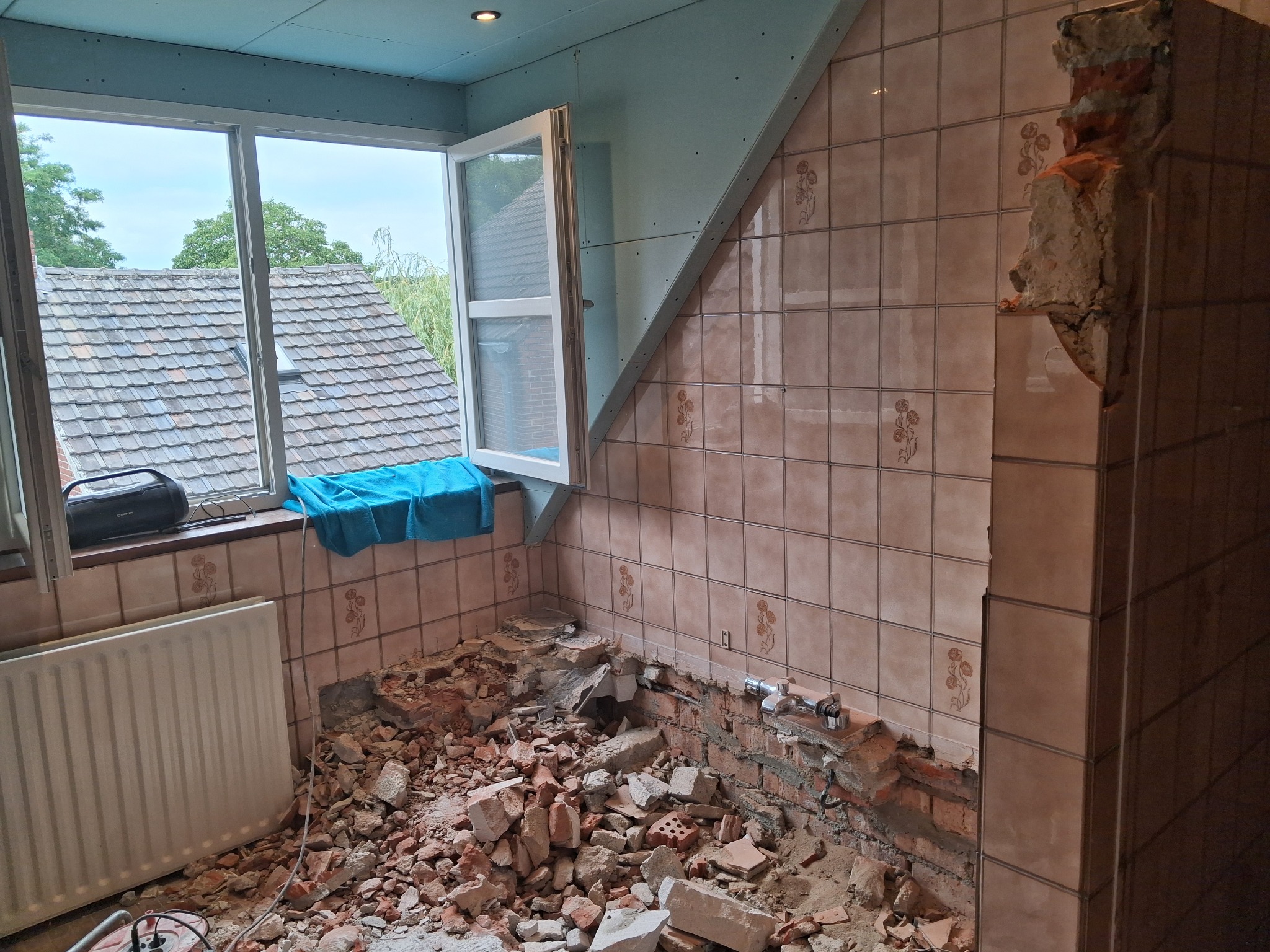 Demontaż płytek w łazience z odsłoniętą instalacją, widoczne gruz i resztki zaprawy. Okno otwarte, na parapecie radio i ręcznik. Ściany częściowo pokryte płytą karton-gips.