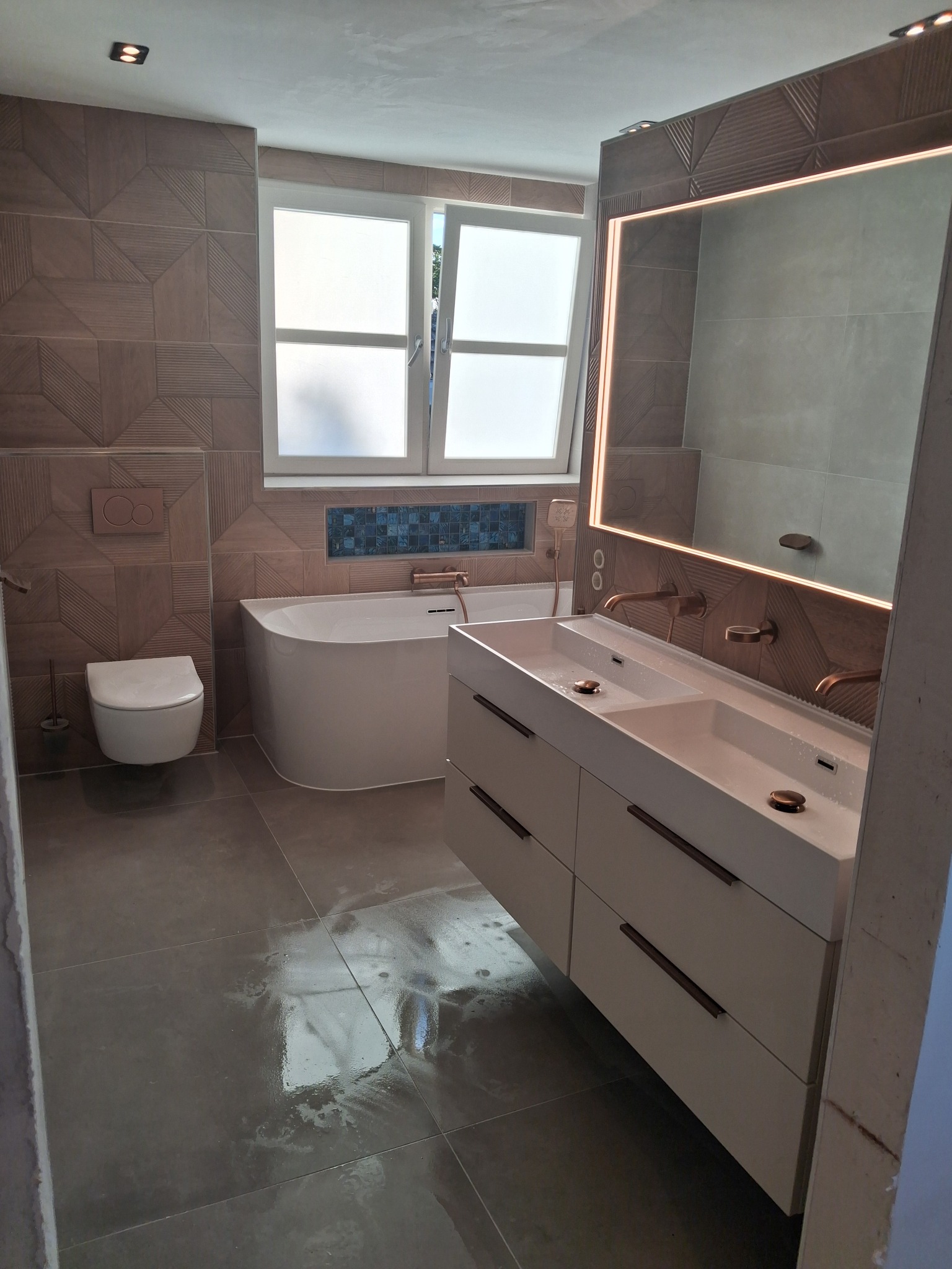 Nowoczesna łazienka po remoncie: brązowe płytki z geometrycznym wzorem, biała wanna i szafka z umywalkami, podświetlane lustro i okno z mleczną szybą.