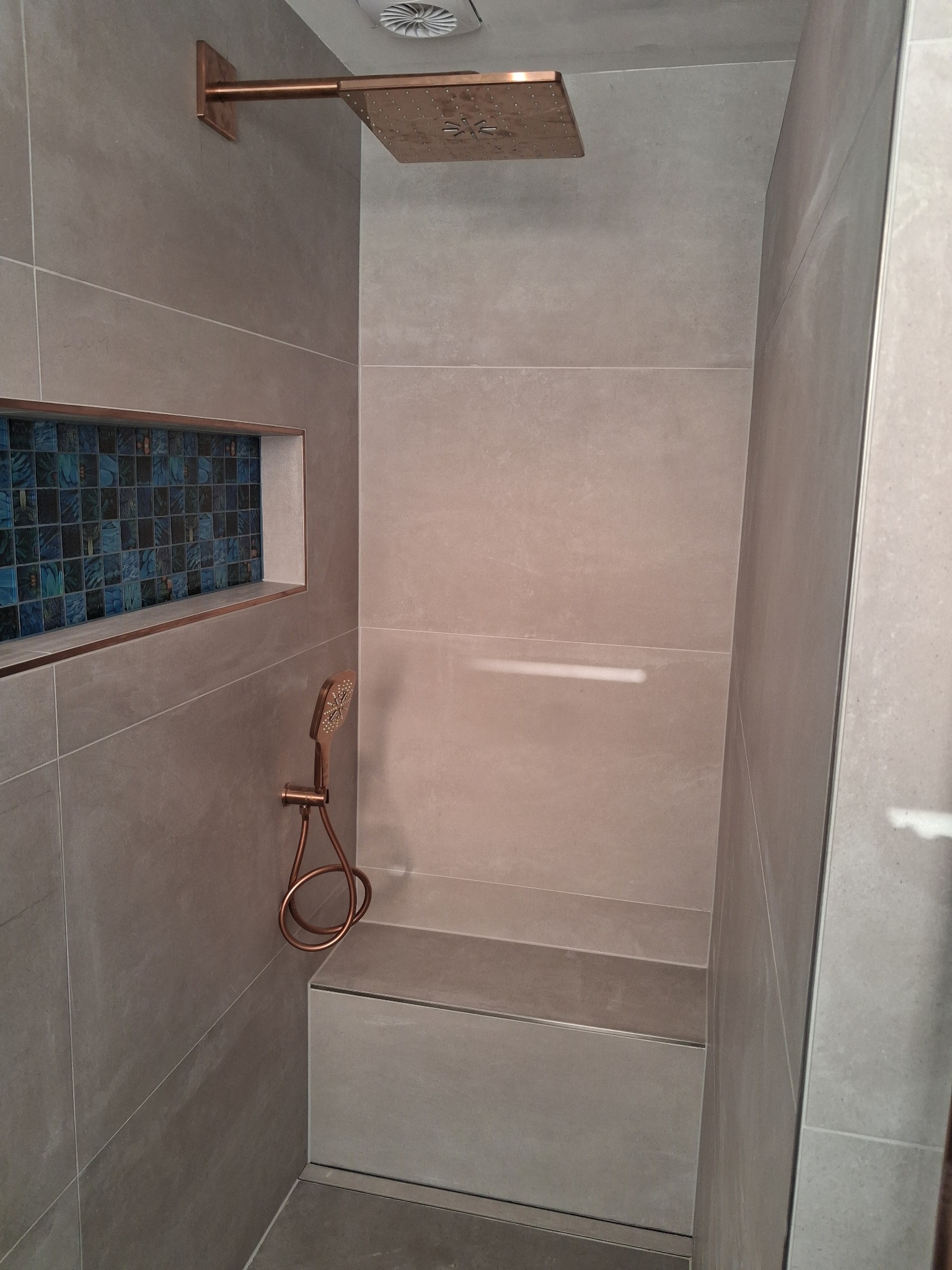 Stylowa kabina prysznicowa z siedziskiem i mozaiką w Berlinie. Szara płytka, miedziana armatura i odpływ liniowy tworzą nowoczesny design łazienki.