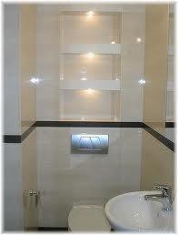 Wykończona łazienka z beżowymi płytkami imitującymi marmur, podświetlana wnęka z półkami, umywalka w kształcie muszli i chromowany przycisk spłuczki.