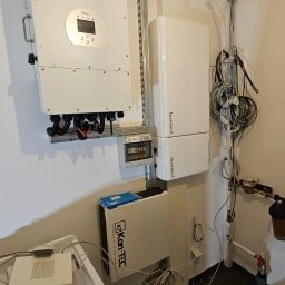 ML SYSTEM+ SPÓŁKA Z OGRANICZONĄ ODPOWIEDZIALNOŚCIĄ - Biały falownik fotowoltaiczny z wyświetlaczem, moduł magazynu energii i skrzynka elektryczna na ścianie wewnątrz pomieszczenia, widoczne przewody i rury.