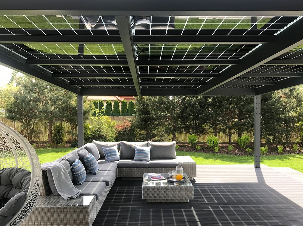 Nowoczesna pergola z panelami słonecznymi na dachu, szary narożnik z poduszkami w paski, szklany stolik kawowy z napojami, wiszące krzesło, zadbany trawnik i drzewa w tle.