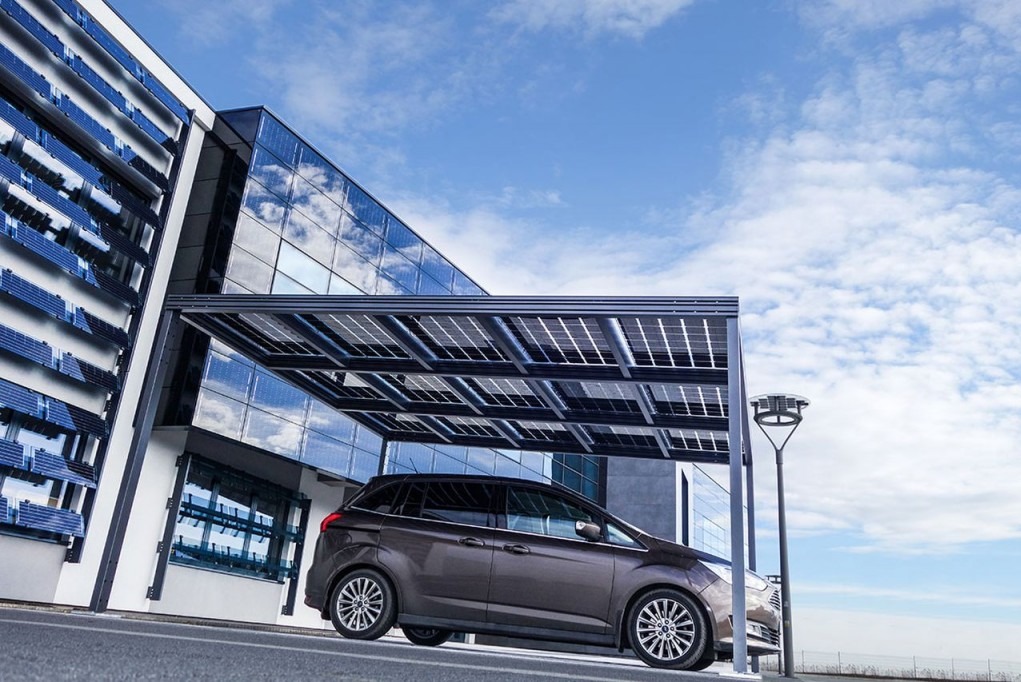 Ciemnobrązowy samochód typu minivan zaparkowany pod carportem z panelami słonecznymi, na tle nowoczesnego budynku z fasadą z paneli fotowoltaicznych i błękitnego nieba z chmurami.