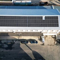 Widok z drona na budynek przemysłowy z zainstalowanymi panelami słonecznymi na dachu. Ciemne panele tworzą regularny wzór na tle jasnoszarego dachu. Na dole kadru widoczne zaparkowane samochody...