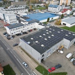 Widok z góry na budynek z panelami słonecznymi na dachu, otoczony innymi budynkami i samochodami, sfotografowany w pochmurny dzień.