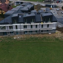 Wielorodzinny budynek mieszkalny w trakcie budowy z panelami słonecznymi na dachu, widok z góry, częściowo ocieplony, z widocznym placem budowy i okoliczną zabudową.