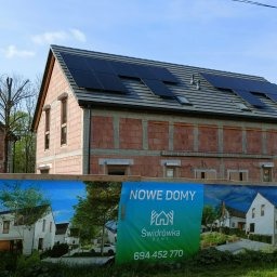 Niedokończony budynek z czerwonej cegły z zainstalowanymi panelami słonecznymi na dachu, widoczne przez baner reklamowy z napisem 'NOWE DOMY'.