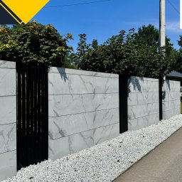 Firma Handlowa PROSPER SYLWIA KUŁAGA-CZERNECKA - Nowoczesne ogrodzenie betonowe z elementami panelowymi w kolorze czarnym, wykończone białym grysem, z logo firmy Prosper w prawym górnym rogu.