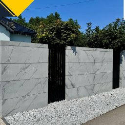 Firma Handlowa PROSPER SYLWIA KUŁAGA-CZERNECKA - Nowoczesne ogrodzenie modułowe z betonowych paneli z marmurkowym wzorem, oddzielone czarnymi, pionowymi elementami, otoczone białym żwirem i zielenią.