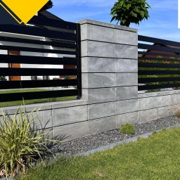 Firma Handlowa PROSPER SYLWIA KUŁAGA-CZERNECKA - Nowoczesne ogrodzenie z betonowych bloczków i poziomych, czarnych paneli, z widocznym logo firmy PROSPER w prawym górnym rogu, otoczone zielenią i czarnym żwirem.