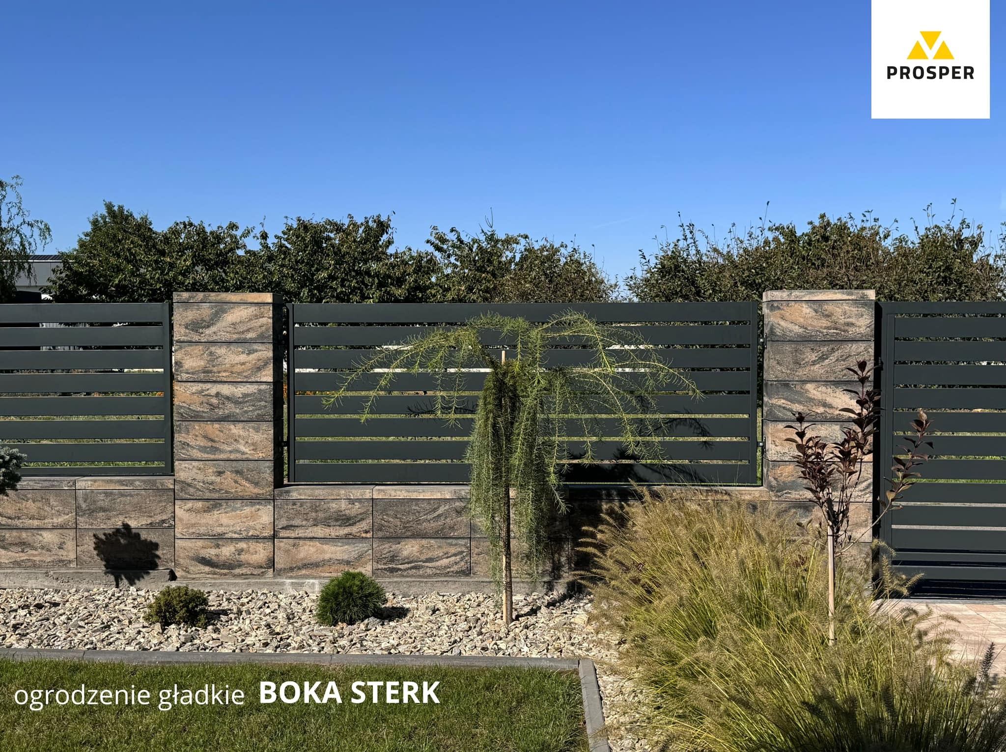 Nowoczesne ogrodzenie z elementami betonowymi imitującymi kamień i poziomymi panelami w kolorze grafitowym, otoczone ozdobnymi krzewami i wysypane drobnym kamieniem, pod błękitnym niebem.