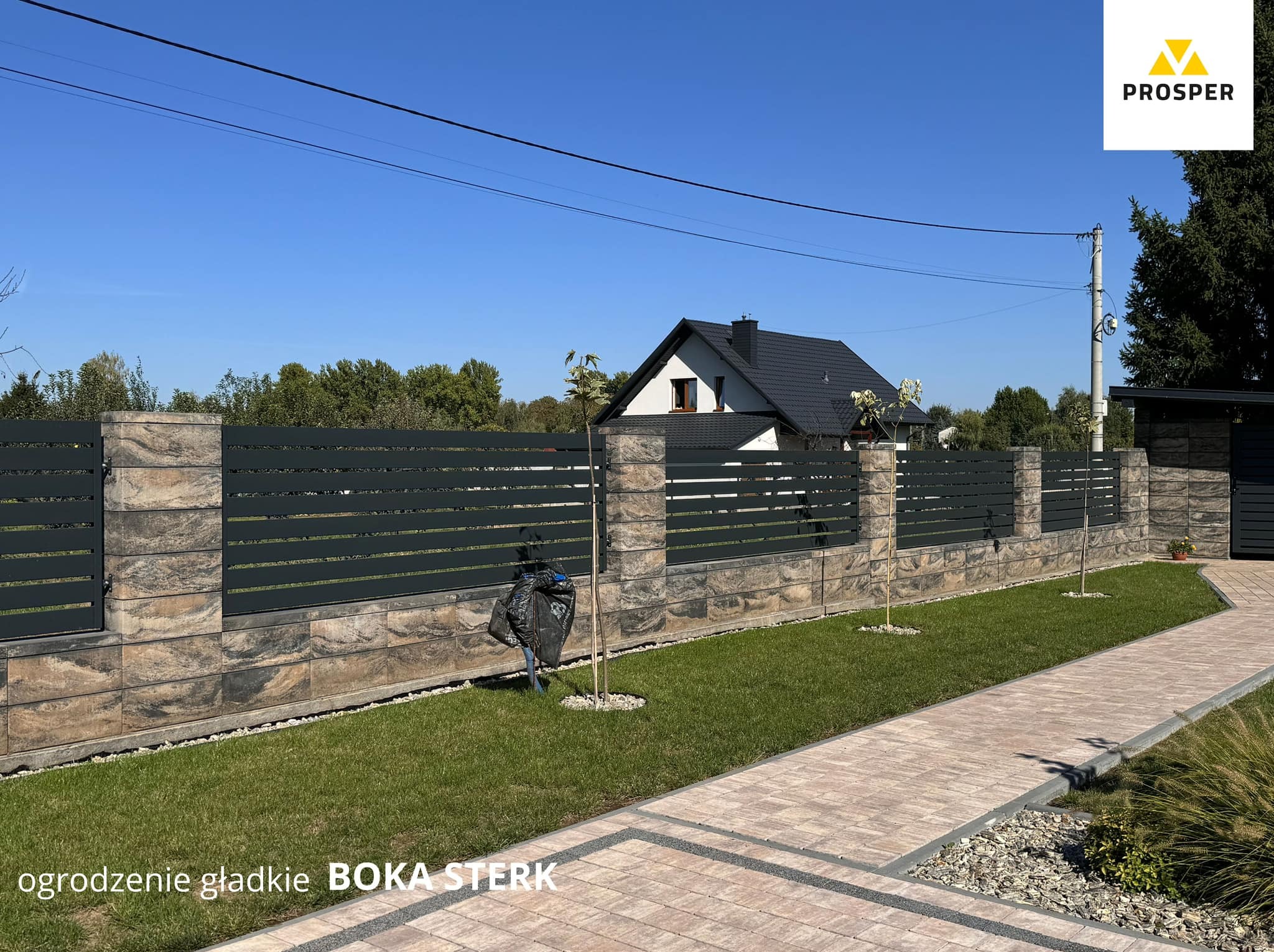 Nowoczesne ogrodzenie z poziomych paneli w kolorze grafitowym, osadzone na słupkach z imitacją kamienia, otoczone zielonym trawnikiem i ścieżką z kostki brukowej, z widocznym domem w tle.