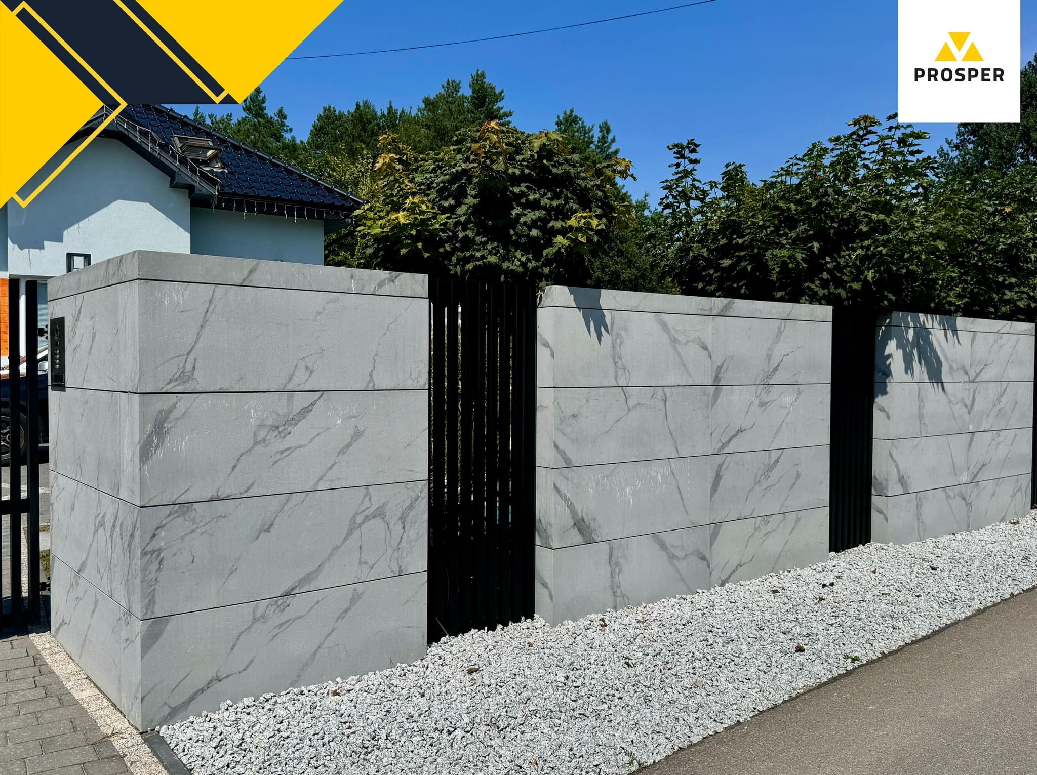 Nowoczesne ogrodzenie modułowe z betonowych paneli z marmurkowym wzorem, oddzielone czarnymi, pionowymi elementami, otoczone białym żwirem i zielenią.
