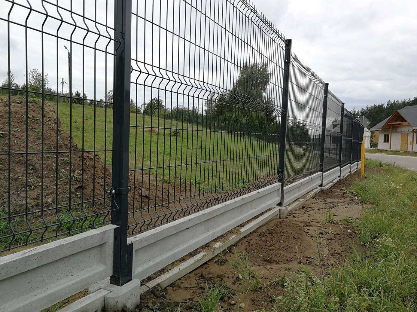 Czarno-szare ogrodzenie panelowe na betonowej podmurówce, widok z boku, trawa i drzewa w tle, fragment drogi z prawej strony.