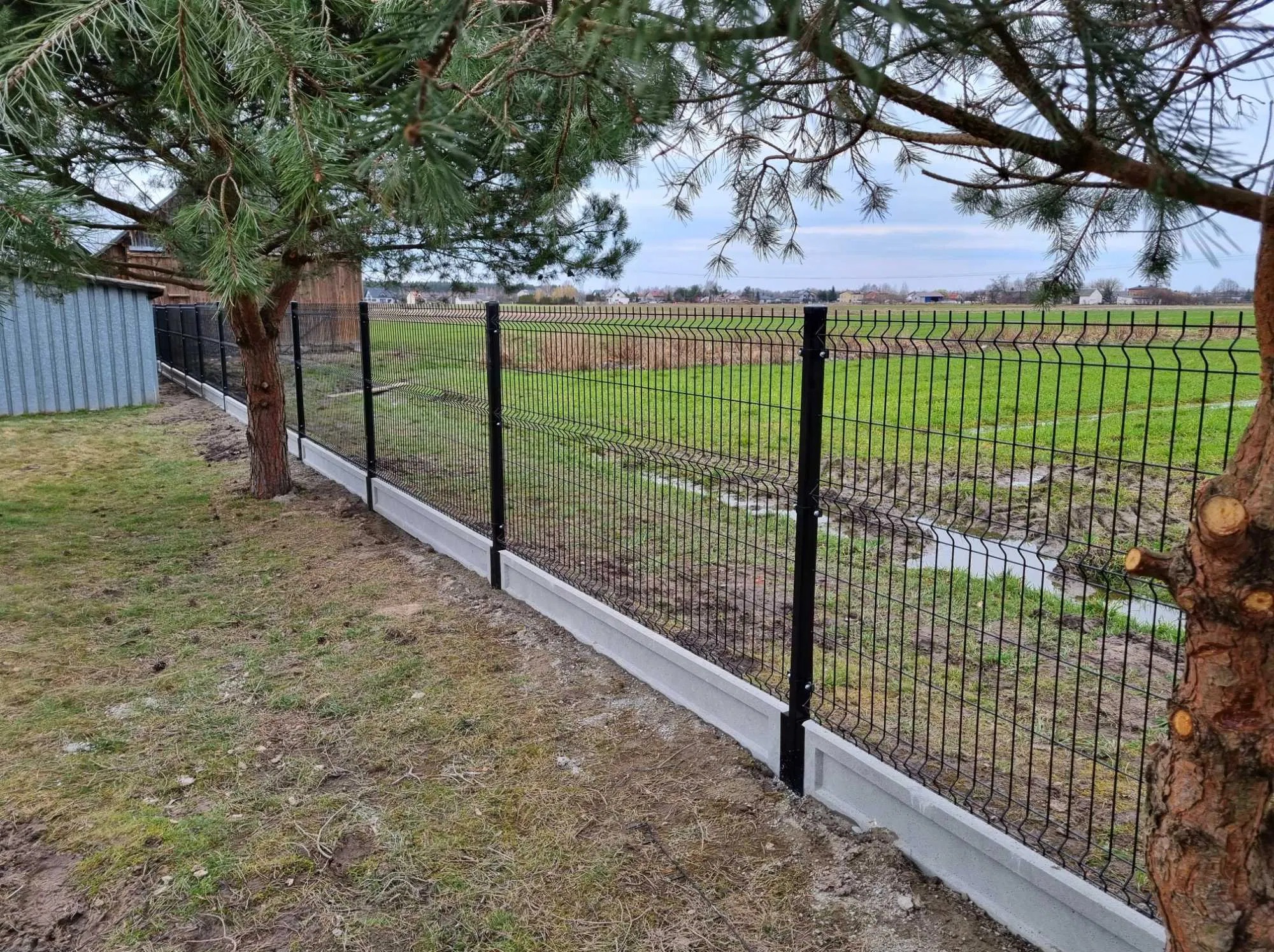 Nowoczesne, czarne ogrodzenie panelowe z podmurówką betonową, widok z perspektywy trawnika z drzewami, w tle łąka i zabudowania.