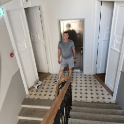 MIPAS-BUD VOLODYMYR PASHCHUK - Wnętrze klatki schodowej z białymi drzwiami z dekoracjami, mozaikową podłogą i mężczyzną stojącym przed windą. Widok z góry na schody z drewnianą barierką.