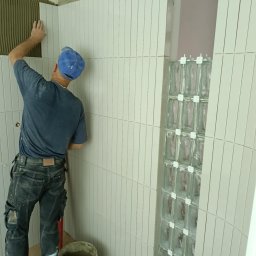 MIPAS-BUD VOLODYMYR PASHCHUK - Układanie pionowych, białych płytek w łazience. Widoczny glazurnik w niebieskiej czapce i fragment ściany z luksferami. Wiadro z zaprawą na podłodze.