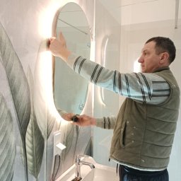 MIPAS-BUD VOLODYMYR PASHCHUK - Mężczyzna montuje owalne lustro z podświetleniem LED w nowoczesnej łazience z tapetą w liście. Widoczna umywalka z baterią i nożyk monterski.