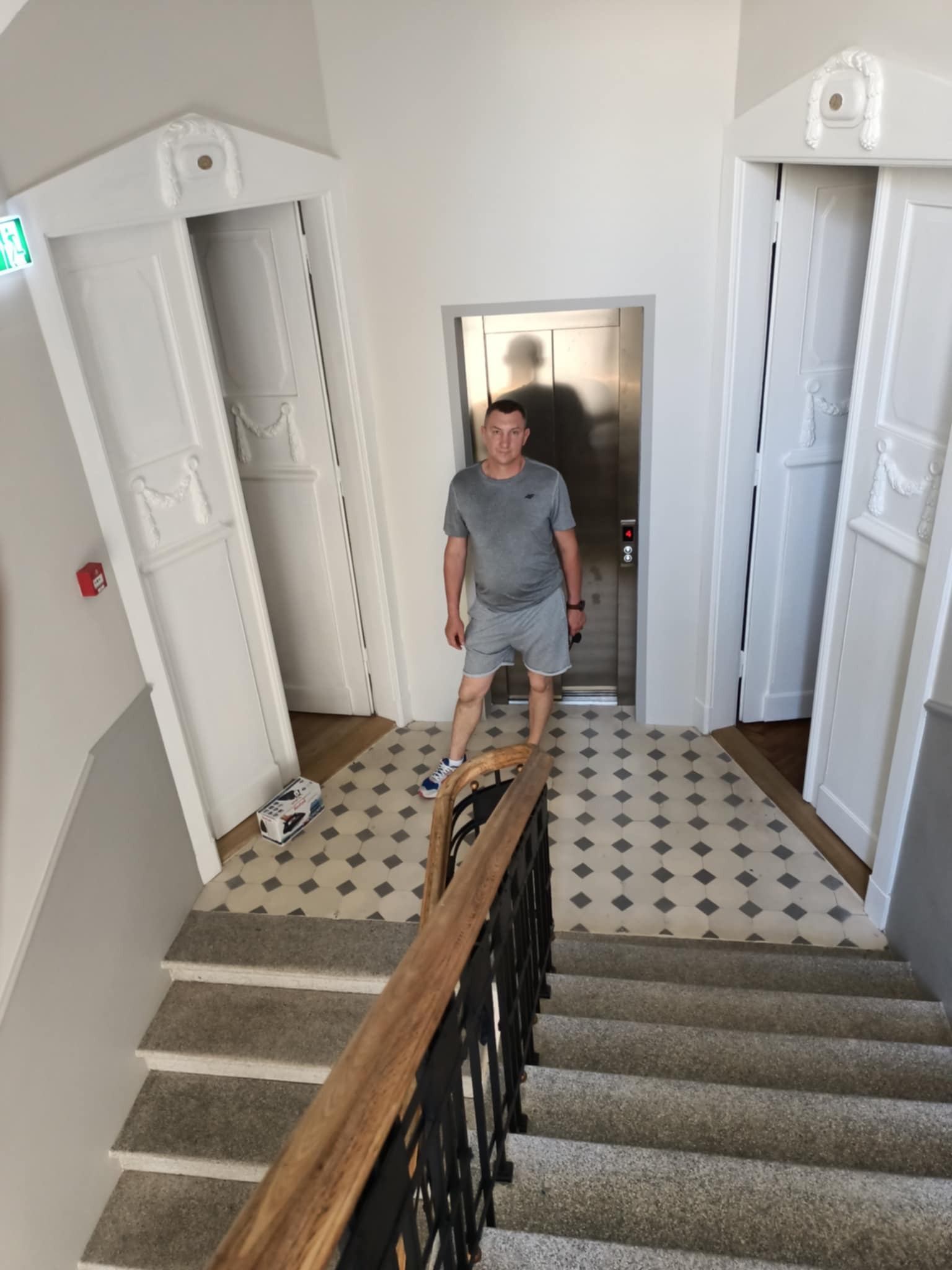 Wnętrze klatki schodowej z białymi drzwiami z dekoracjami, mozaikową podłogą i mężczyzną stojącym przed windą. Widok z góry na schody z drewnianą barierką.