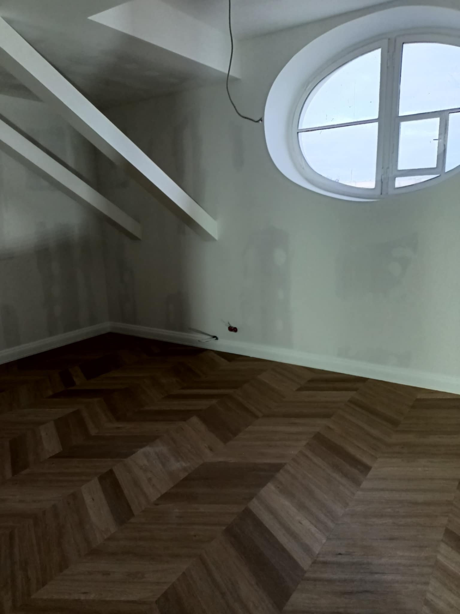 Wnętrze pokoju z parkietem ułożonym we wzór jodełki. Ściany przygotowane do malowania, widoczne belki stropowe i okrągłe okno.