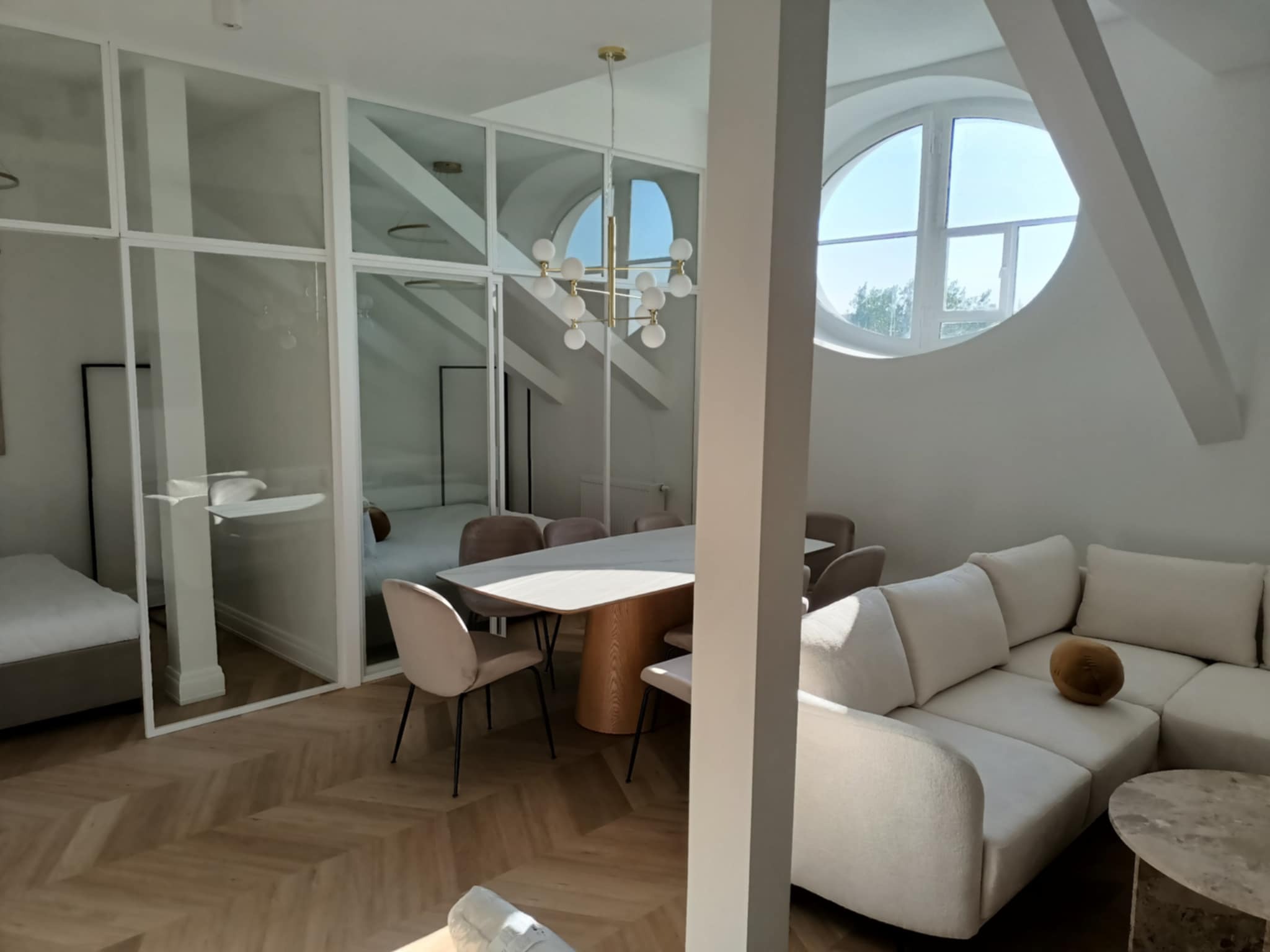 Nowoczesne wnętrze: jasny salon z parkietem w jodełkę, szklane ściany działowe, okrągłe okno na poddaszu i designerska lampa nad stołem. Minimalistyczny wystrój.