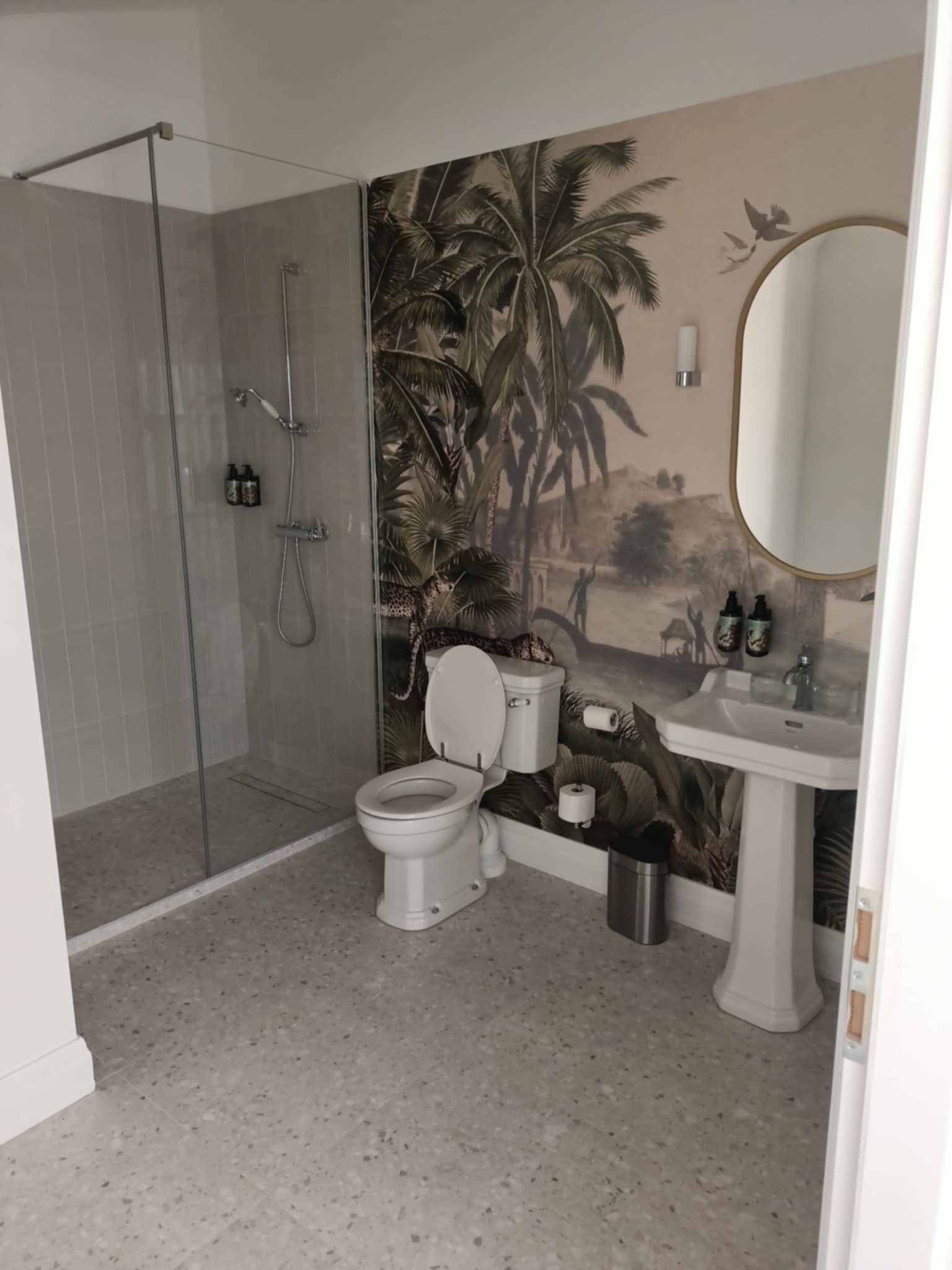 Jasna łazienka z kabiną prysznicową i umywalką na postumencie. Na ścianie tapeta w motywy roślinne i zwierzęce. Lustro w złotej ramie. Całość utrzymana w stonowanej kolorystyce.