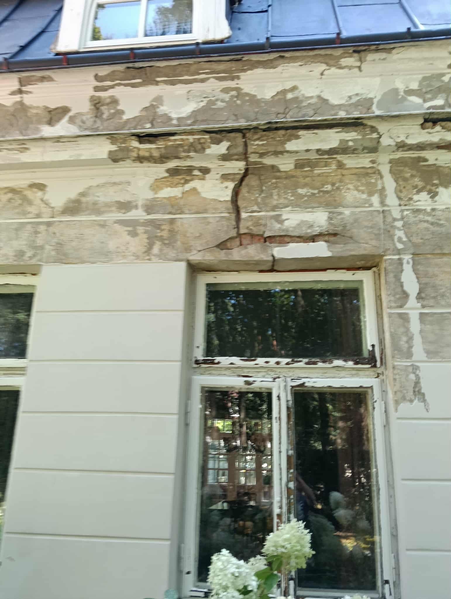 Zniszczona elewacja budynku z odpadającym tynkiem i pęknięciem nad oknem. Widoczne fragmenty muru i stara stolarka okienna. Dolny kadr z kwiatami hortensji.