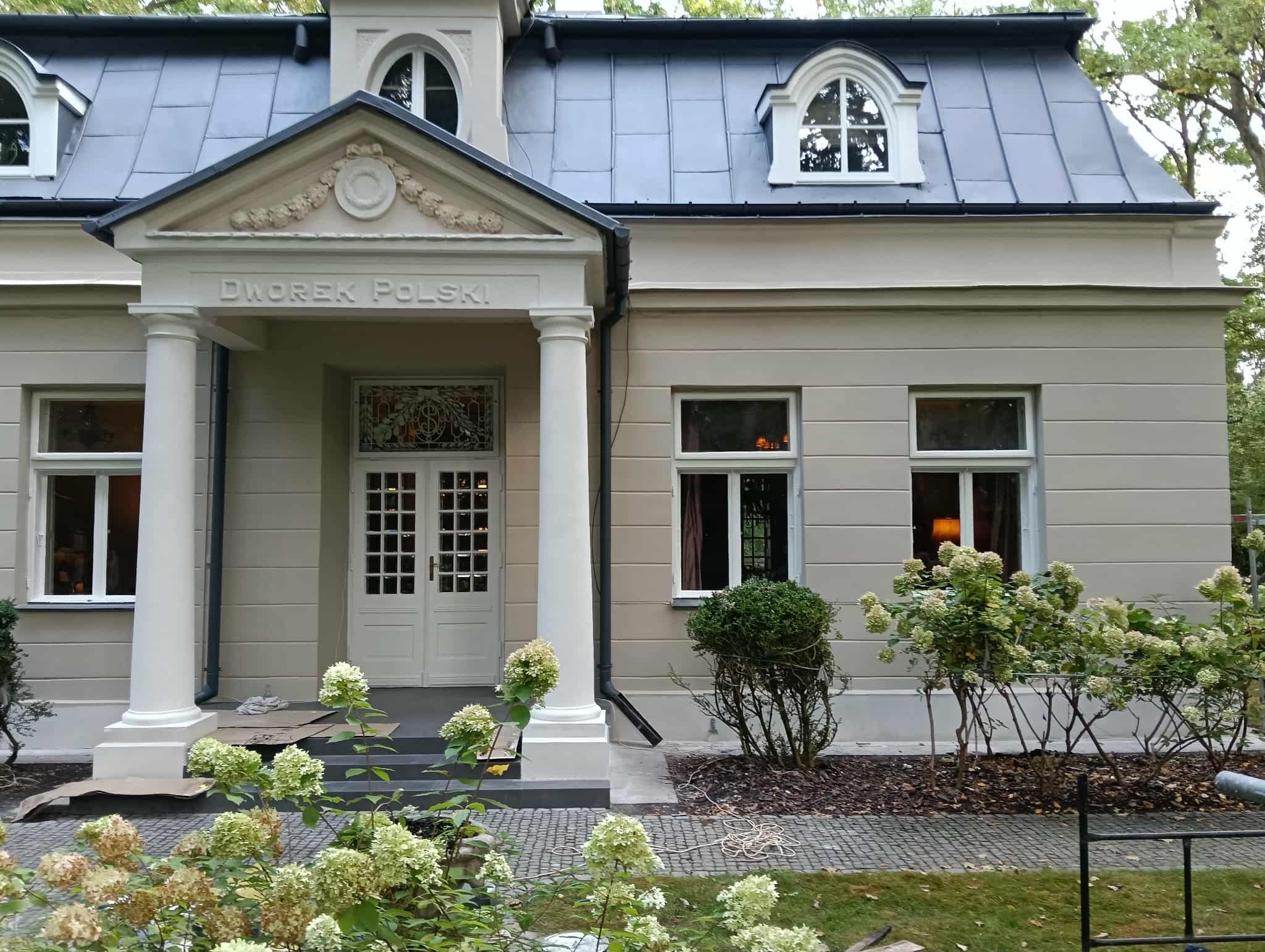 Odnowiony dworek z kolumnami i napisem 'Dworek Polski' nad wejściem, otoczony kwitnącymi hortensjami. Widoczne świeżo pomalowane elementy fasady.