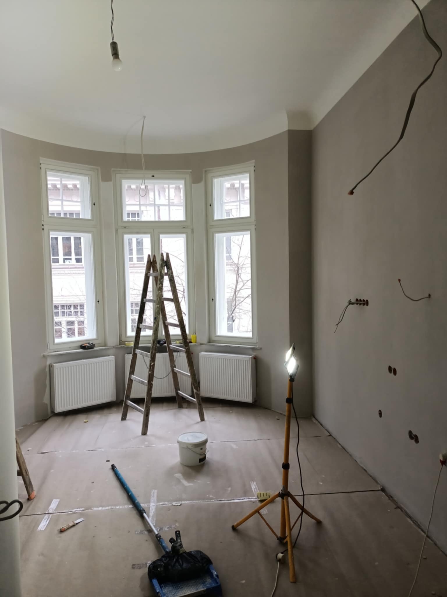 Pokój w trakcie malowania: drabina, wiadro z farbą, wałek i lampa budowlana. Ściany częściowo pomalowane na szaro, widoczne instalacje elektryczne.