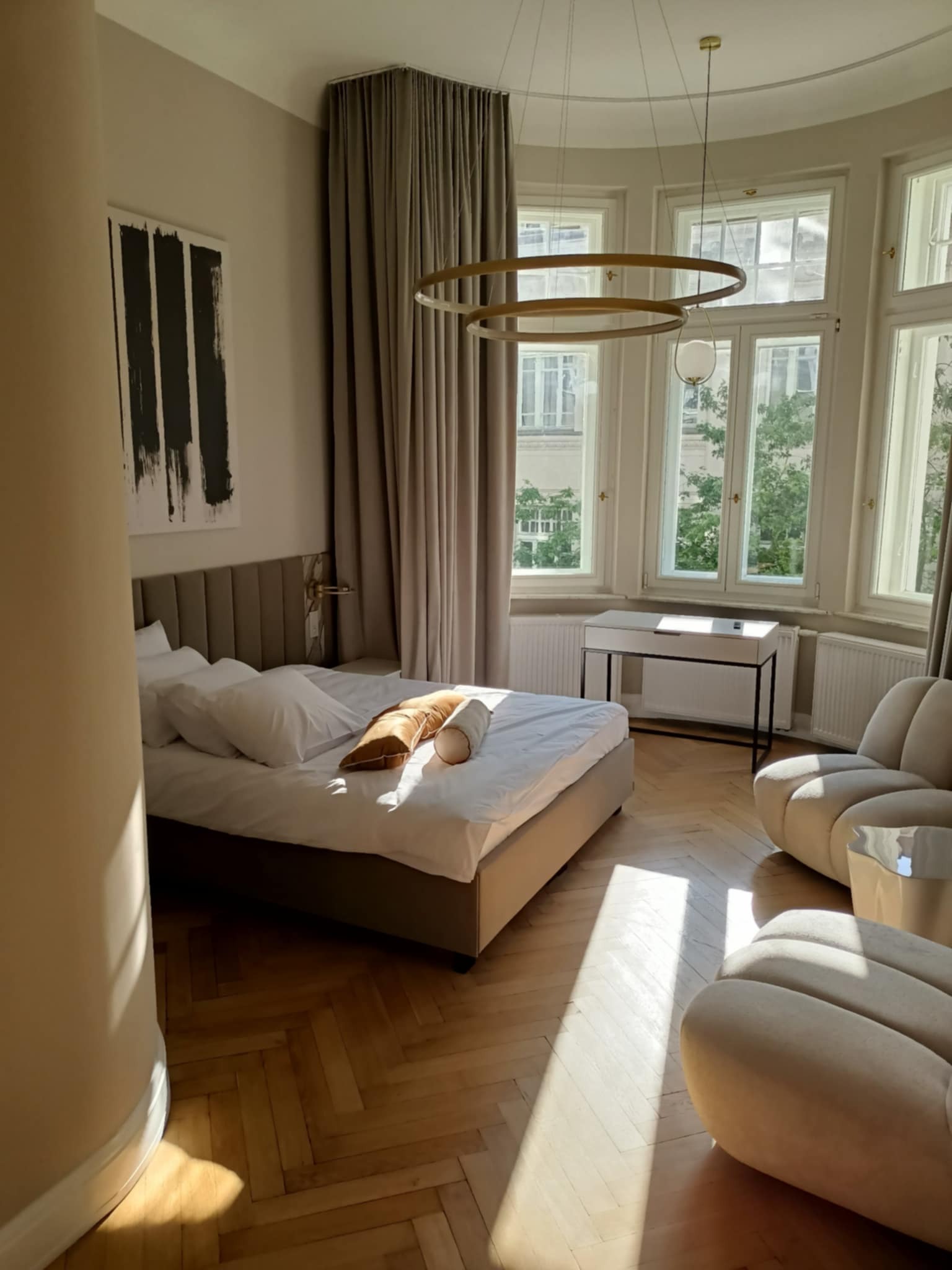 Jasna sypialnia w Uwielinach z parkietem w jodełkę, beżowymi zasłonami i designerską lampą. Duże okna wpuszczają promienie słoneczne.