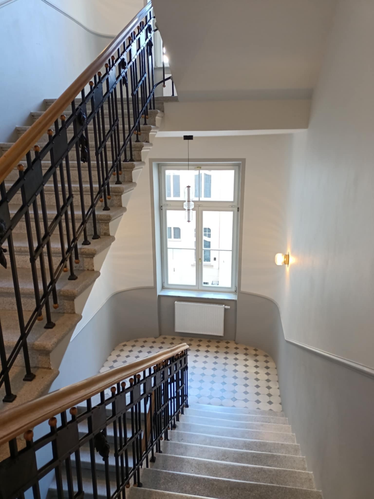 Klatka schodowa w kamienicy po remoncie: szare ściany, granitowe schody z czarną balustradą, okno z grzejnikiem i wzorzysta podłoga na podeście. Widok z góry.