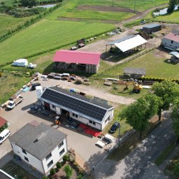 ASTA ENERGY SYSTEMS SPÓŁKA Z OGRANICZONĄ ODPOWIEDZIALNOŚCIĄ - Widok z lotu ptaka na budynek z panelami słonecznymi na dachu, otoczony zabudowaniami gospodarczymi i polami uprawnymi w słoneczny dzień.