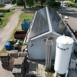 ASTA ENERGY SYSTEMS SPÓŁKA Z OGRANICZONĄ ODPOWIEDZIALNOŚCIĄ - Instalacja paneli na dachu budynku przemysłowego, widok z góry. Obok zbiornik Messer i palety. W tle plac z koparką i kontenerami.