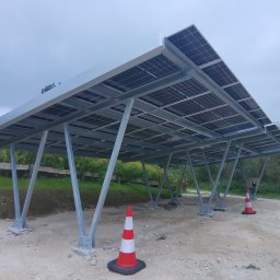 ASTA ENERGY SYSTEMS SPÓŁKA Z OGRANICZONĄ ODPOWIEDZIALNOŚCIĄ - Zadaszenie parkingowe z panelami słonecznymi, konstrukcja stalowa, widoczne słupki i panele, teren budowy z pachołkami. Pochmurne niebo w tle.