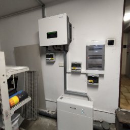 ASTA ENERGY SYSTEMS SPÓŁKA Z OGRANICZONĄ ODPOWIEDZIALNOŚCIĄ - Biały inwerter Sofar i akumulator energii w pomieszczeniu, z widoczną rozdzielnicą elektryczną i przewodami. Instalacja fotowoltaiczna w garażu.