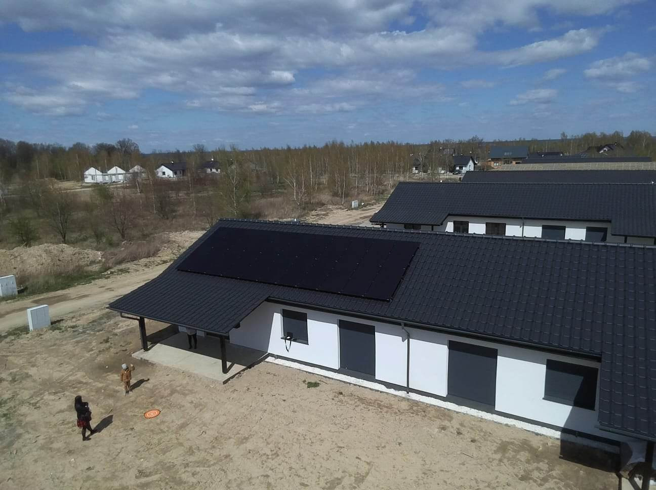 Nowoczesny dom jednorodzinny z panelami słonecznymi na dachu, widok z góry. Biała elewacja, grafitowa dachówka i stolarka okienna. W tle zabudowa jednorodzinna i las.