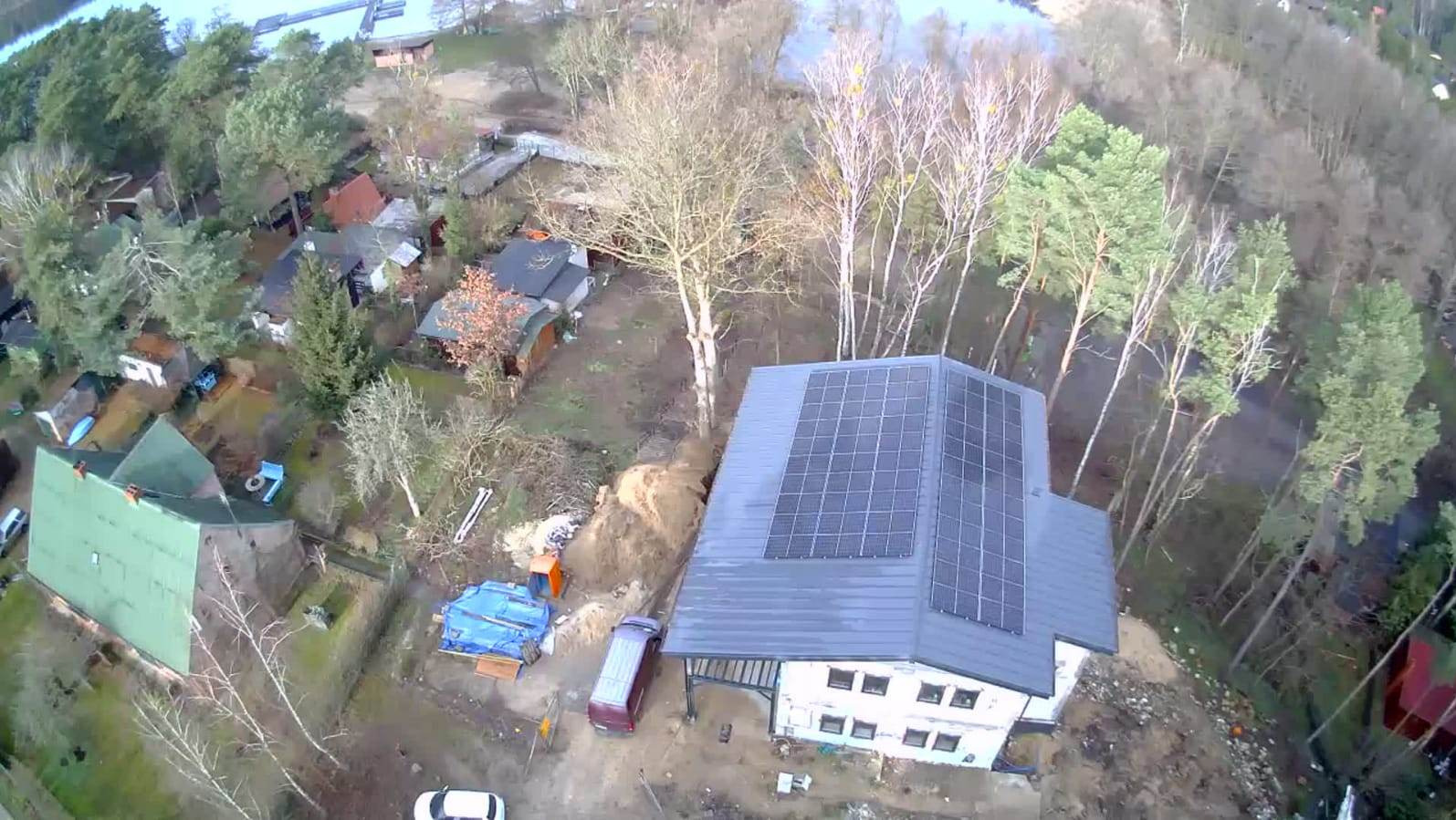 Dom z panelami słonecznymi na dachu, widok z drona. W tle drzewa i jezioro. Na podwórku zaparkowany samochód dostawczy i sterta piasku.