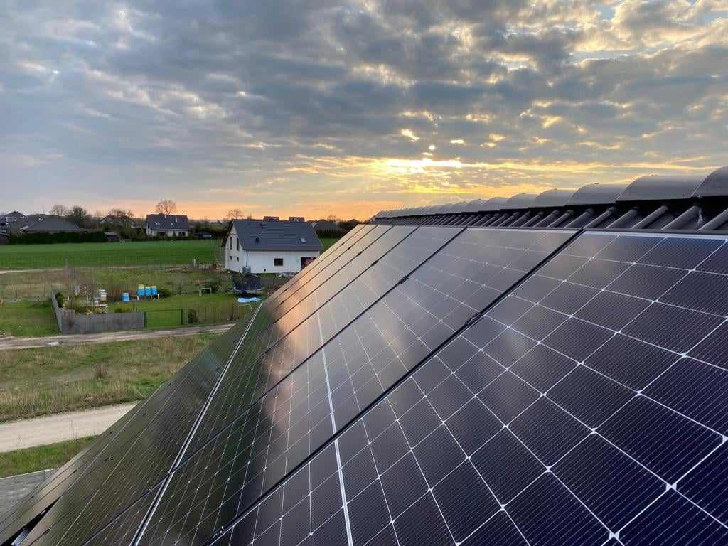 Panele słoneczne na dachu domu jednorodzinnego, widok z góry, w tle zachodzące słońce i zabudowa wiejska. Instalacja fotowoltaiczna w promieniach zachodzącego słońca.